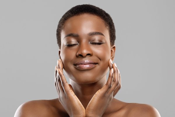 Perfect skin. Portrait of beautiful african american woman with natural makeup touching her face with pleased face expression over grey background